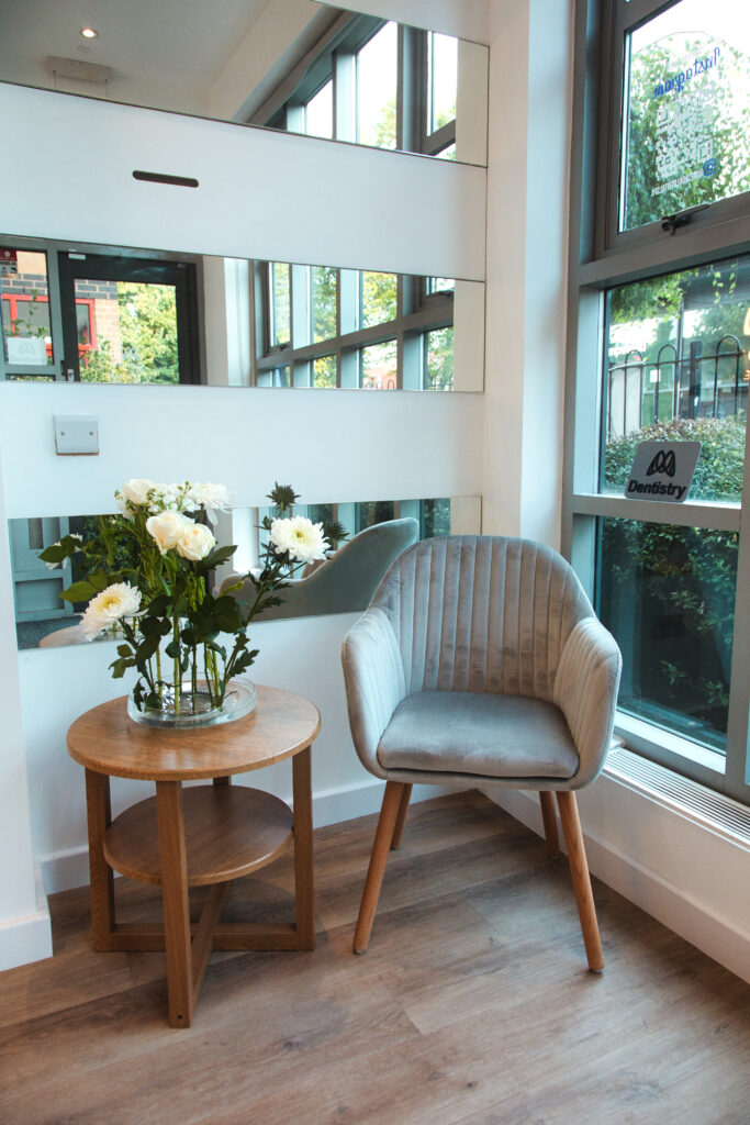 Cozy seating corner at Medical Dental Spa with a modern grey chair, wooden side table, and fresh flowers by large windows.