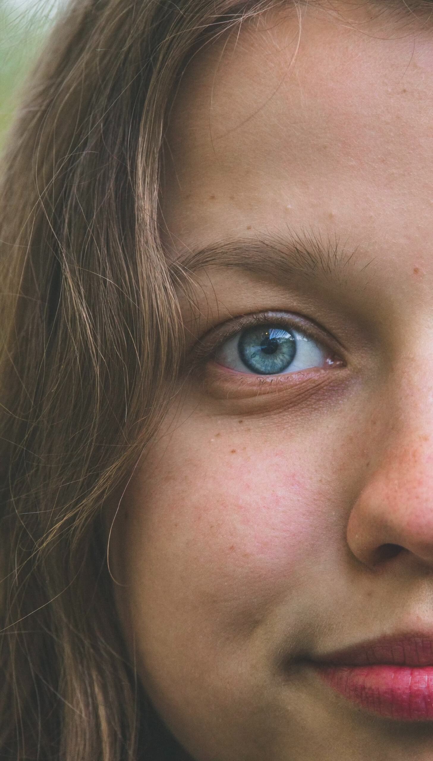 Close-up of a young girl with blond hair, blue eyes and red lips. Only half of the face is visible.