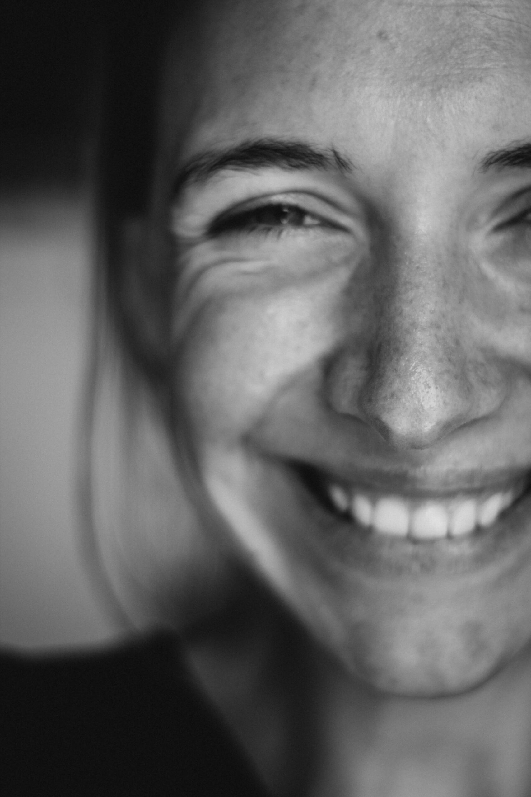 B&W close-up photo of a younger woman with a nig smile, wrinkles around the eye. the background is out of focus.