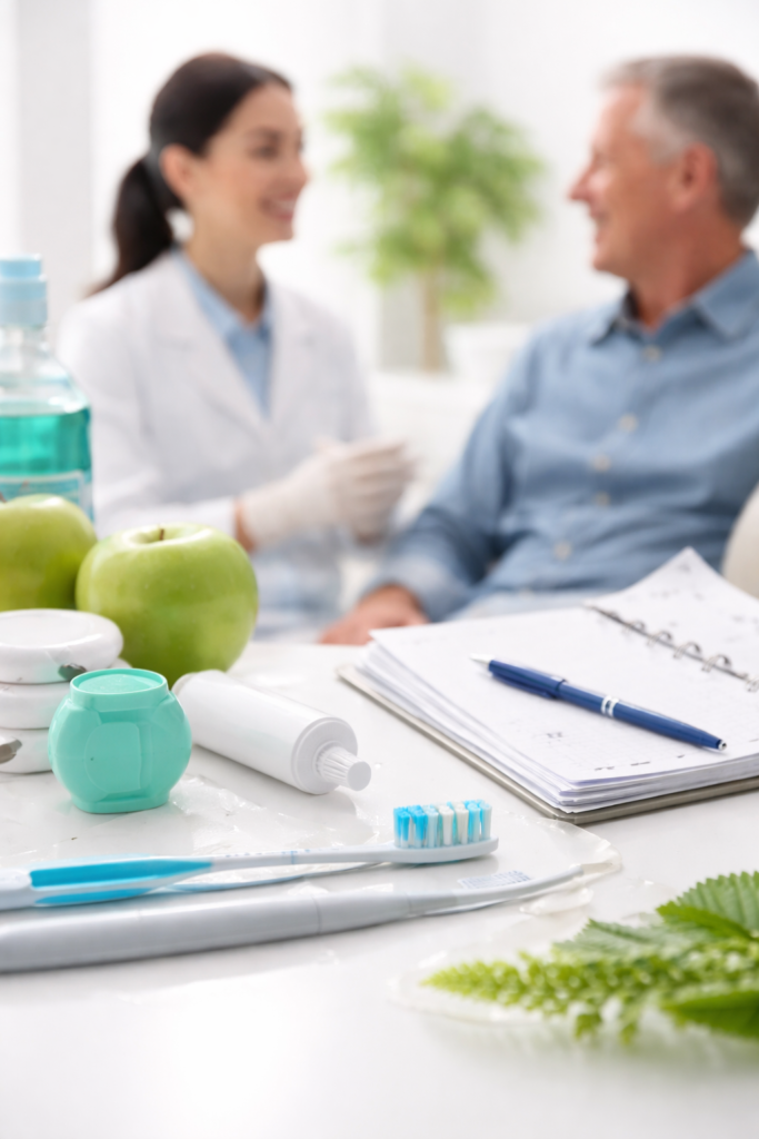Bright, calming dental clinic scene showing a toothbrush, toothpaste, floss, and fresh apples arranged on a clean surface in the foreground, with a dentist warmly talking to a relaxed patient in the softly blurred background, representing preventive oral care and overall wellbeing.